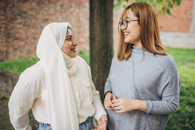 An Asian female talks with a Muslim friend while standing in backyard outdoors.